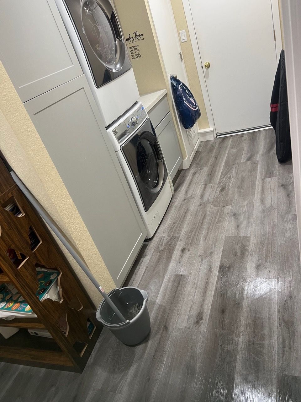 Laundry room with stacked washer and dryer, gray floor, and cleaning supplies near the wall.
