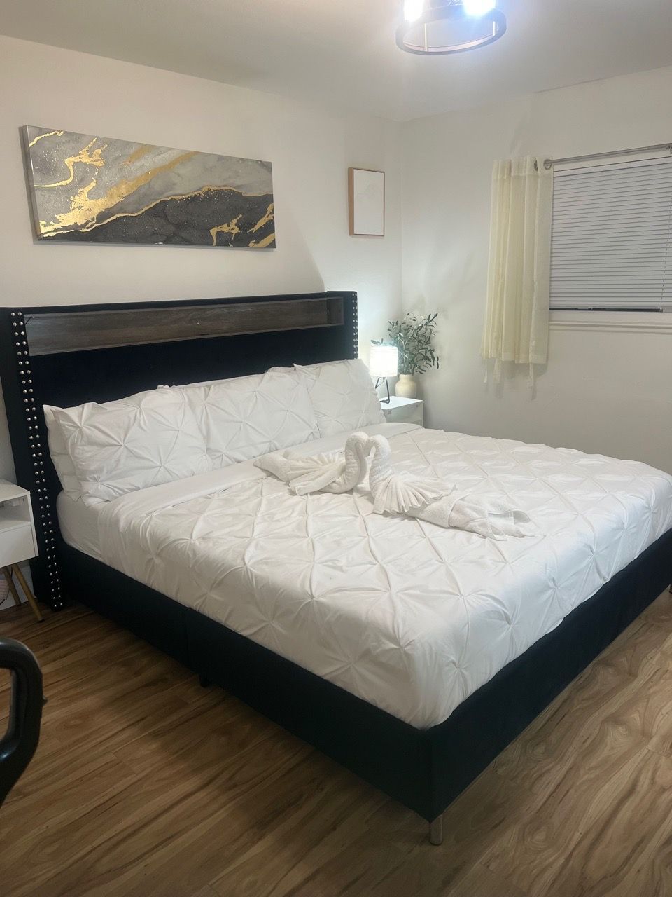 Neatly made bed in a bright bedroom with white bedding, black headboard, and wood flooring.
