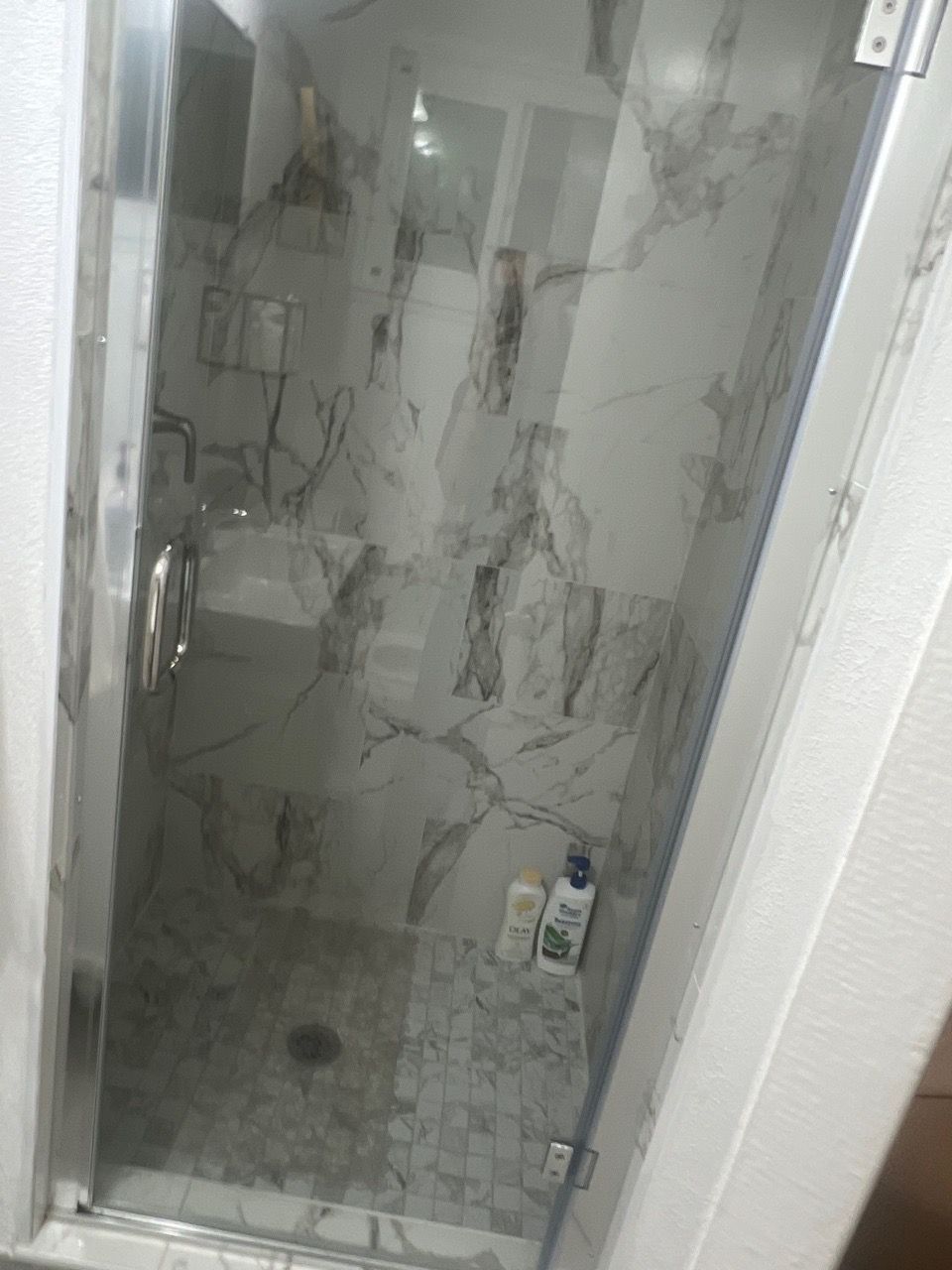 Small tiled shower stall with glass door, stone-pattern walls, and a bottle on the floor.