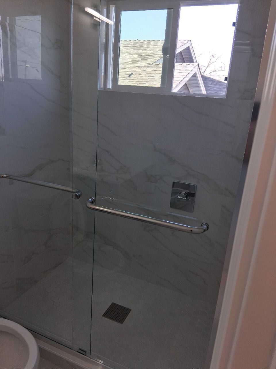 Glass shower stall with chrome grab bars, white tile walls, and a small window above the shower area