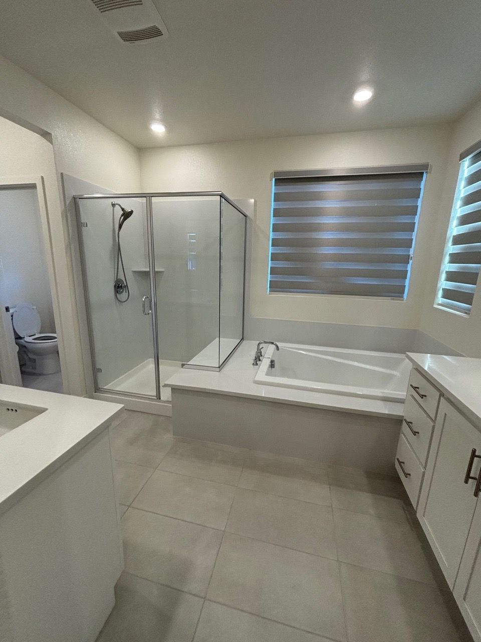 Modern bathroom with a glass shower, large soaking tub, double vanity, and window blinds.