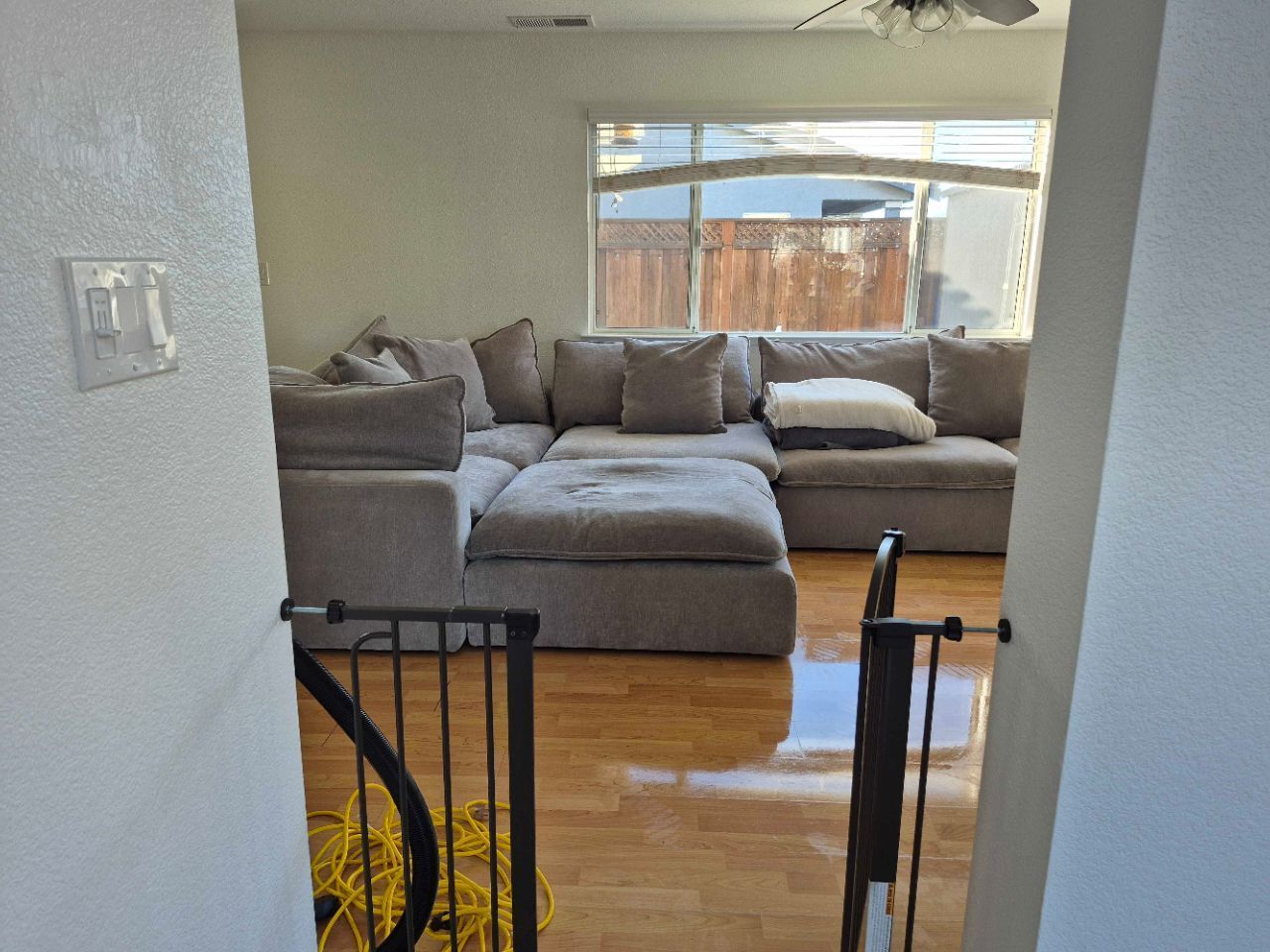 Cozy living room with gray sectional sofa, hardwood floor, and large window overlooking a fenced yard