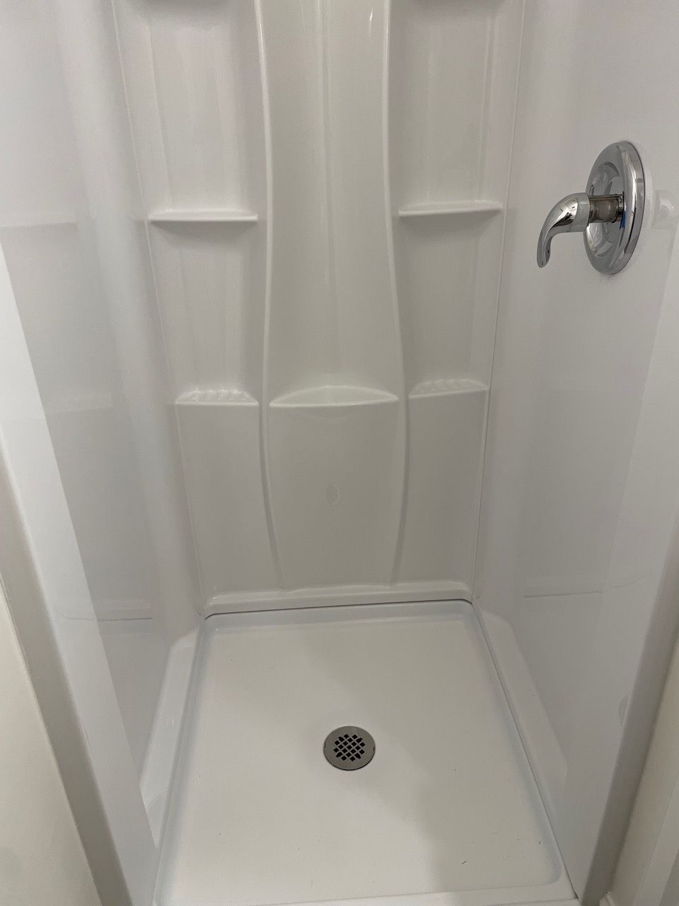 White shower stall with built-in shelves, chrome shower handle, and drain.
