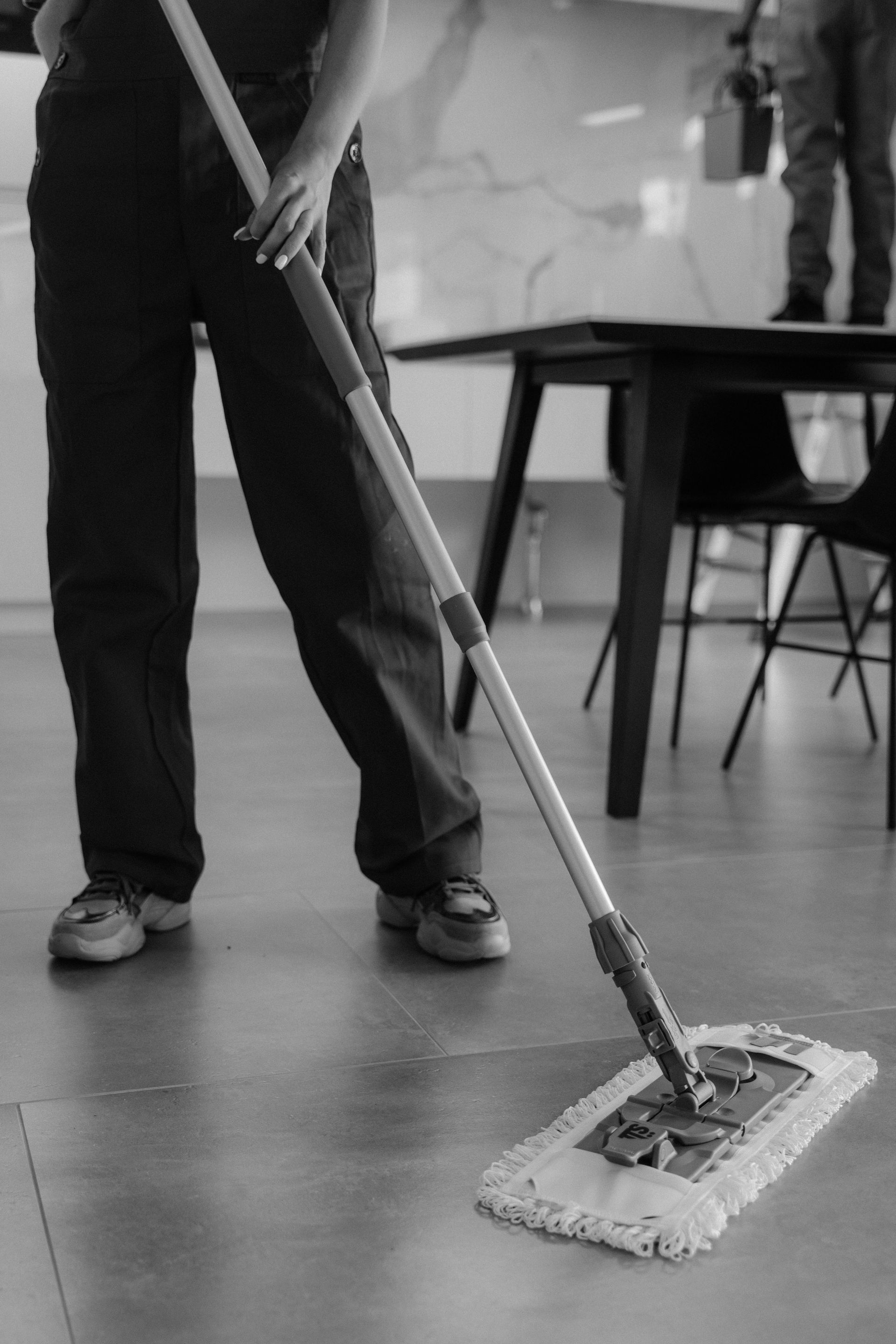 Person mopping a floor in a room with chairs and tables nearby.