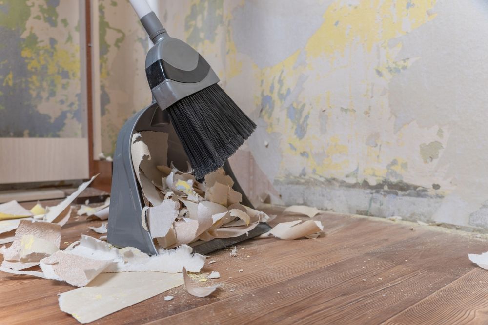 Dustpan and broom sweeping up broken white plaster on a wood floor near a damaged wall.