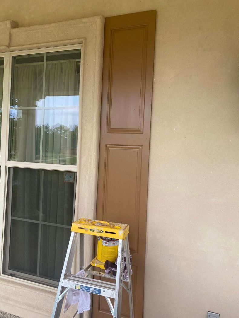 A step ladder holds a brown shutter panel beside a window on a stucco exterior wall.