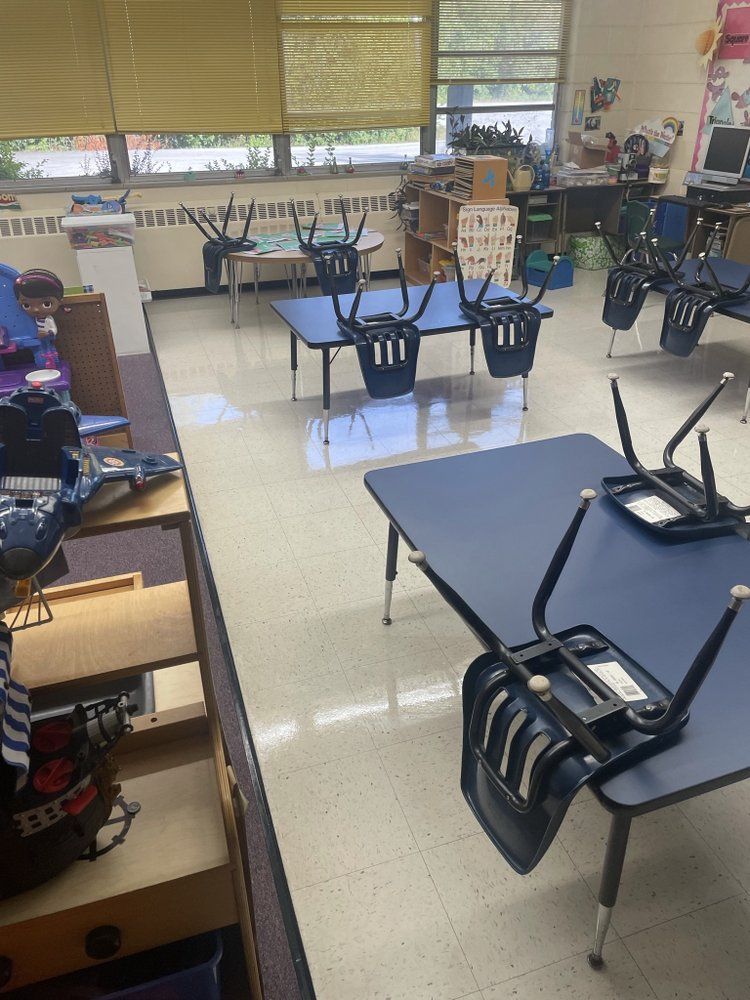 Empty classroom with desks, chairs upside down, and windows with blinds along the back wall.