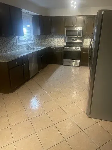 Empty modern kitchen with dark cabinets, beige tile floor, stainless steel appliances, and a window over the sink