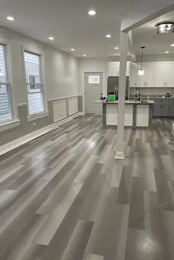 Empty modern kitchen and living area with gray wood floors, white walls, and recessed lighting
