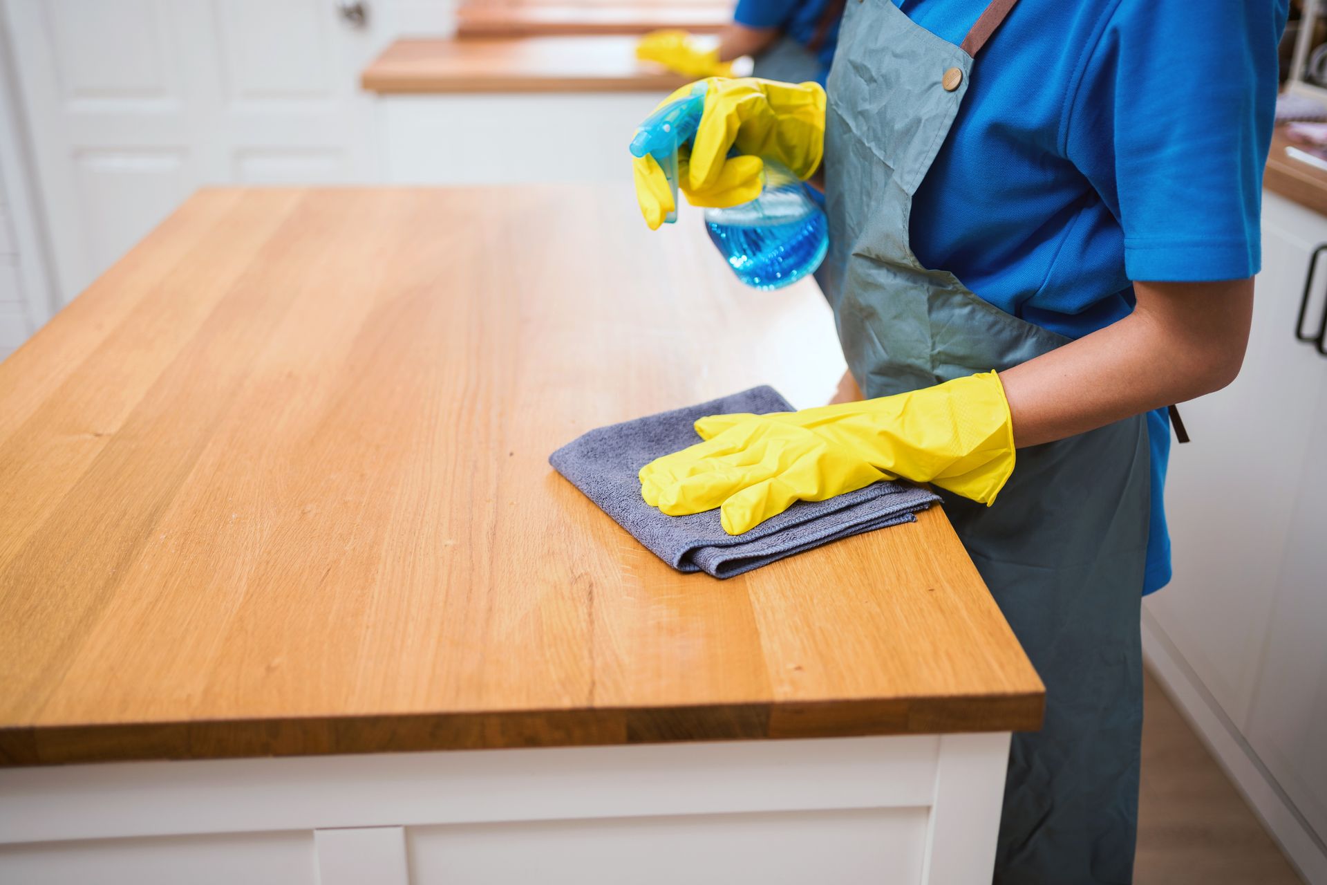 Person cleaning a wooden table with spray and cloth, wearing yellow gloves and a blue shirt