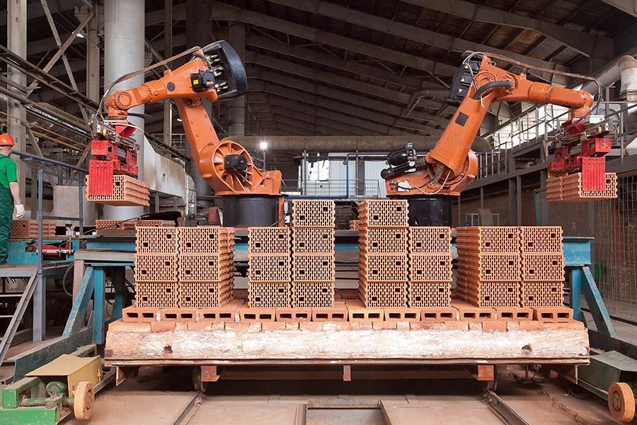 Due bracci robotici arancioni impilano mattoni su pallet all'interno di un magazzino industriale.