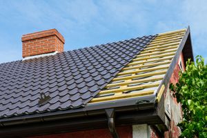Roof under repair with exposed yellow sheathing beside dark gray shingles and a brick chimney