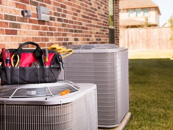 Two outdoor HVAC units beside a brick house, with a tool bag resting on the front unit.