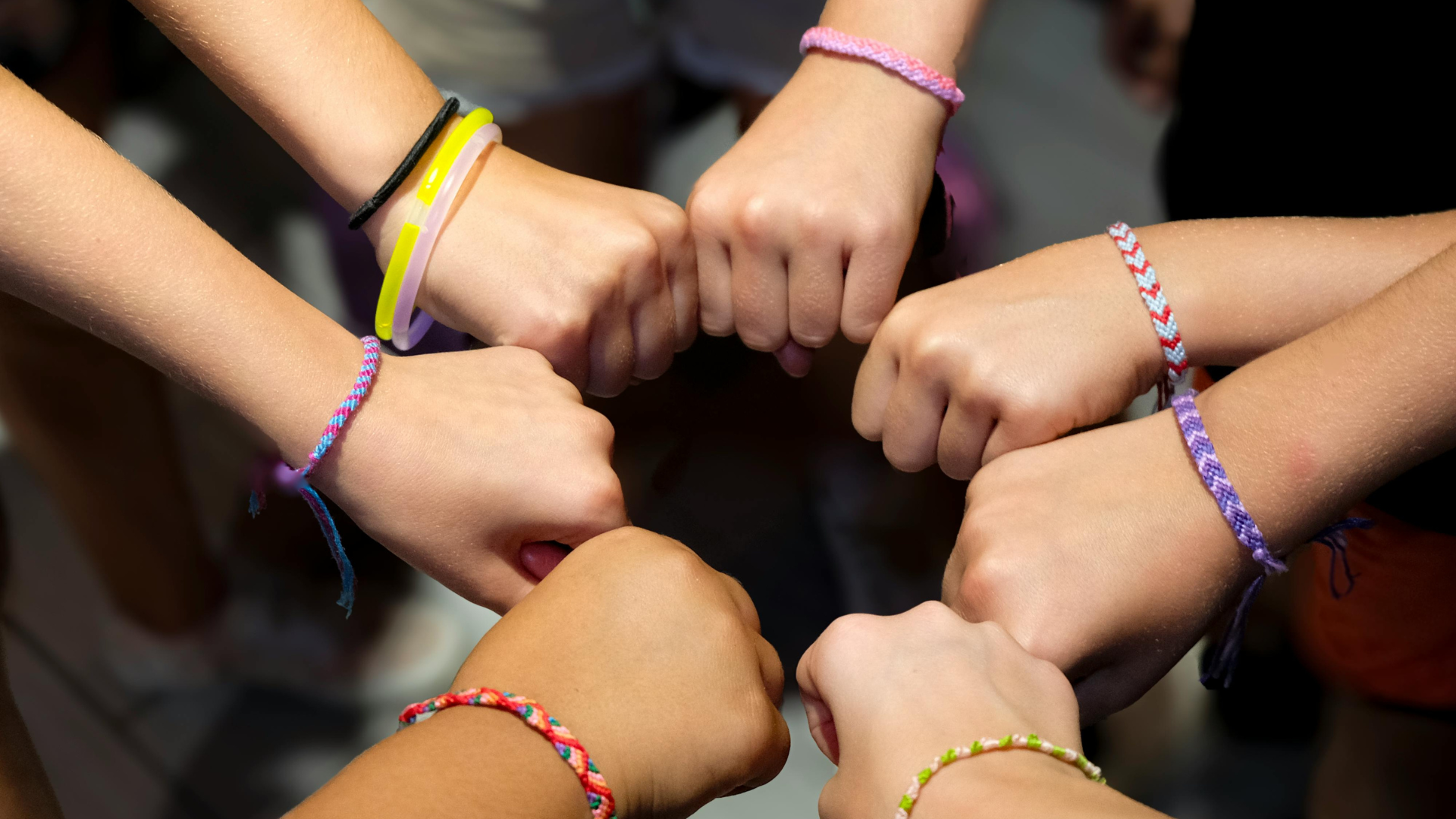Six fists stacked together in a circle, each wearing colorful bracelets.