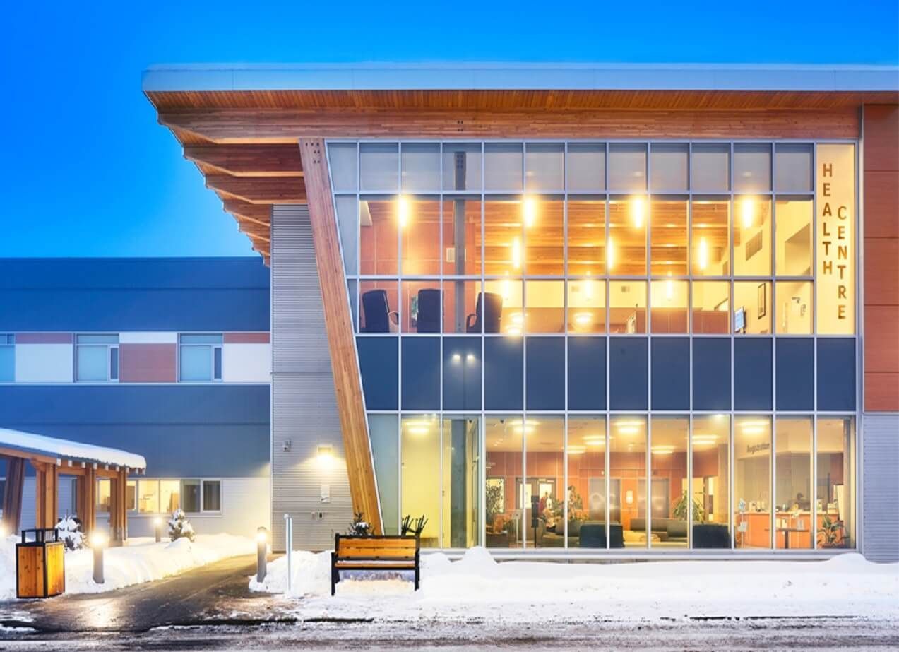 Health center entrance, snow-covered, large windows, wood accents, and evening lighting.