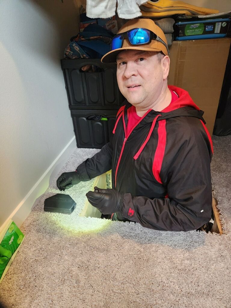 Person in a black-and-red jacket and cap kneels in a carpeted closet, shining a flashlight near storage bins.
