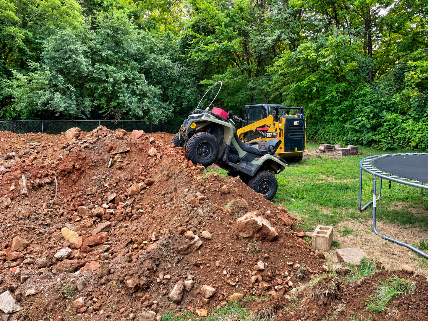 Yellow ATV on a dirt mound near a trampoline in a wooded yard