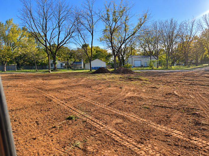 Freshly graded dirt lot with trees and small white buildings under a clear blue sky