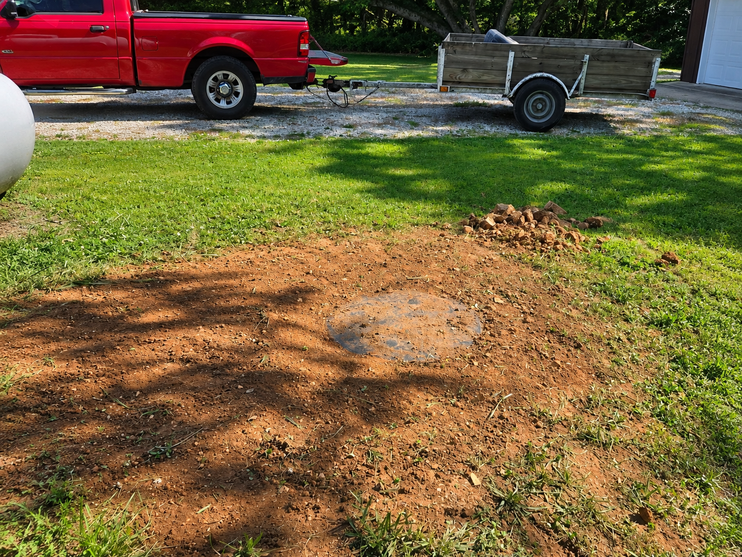 Dirt pile in a grassy yard with a red pickup truck and small trailer in the background