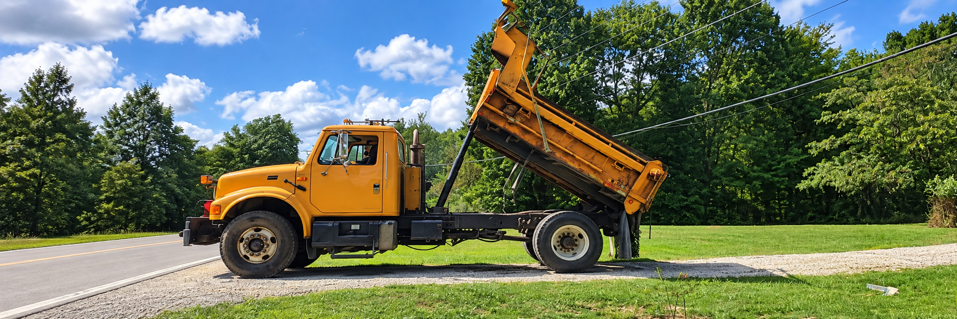 Orange dump truck with raised bed on a roadside beside green grass and trees under a blue sky