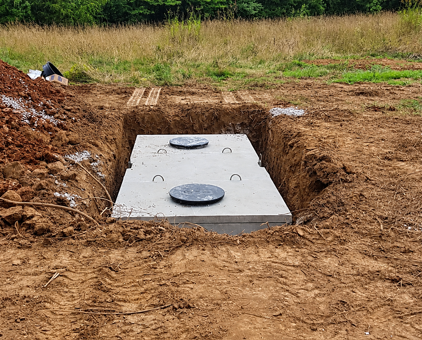 Concrete septic tank lid in a dirt trench at a construction site, with two round access covers visible