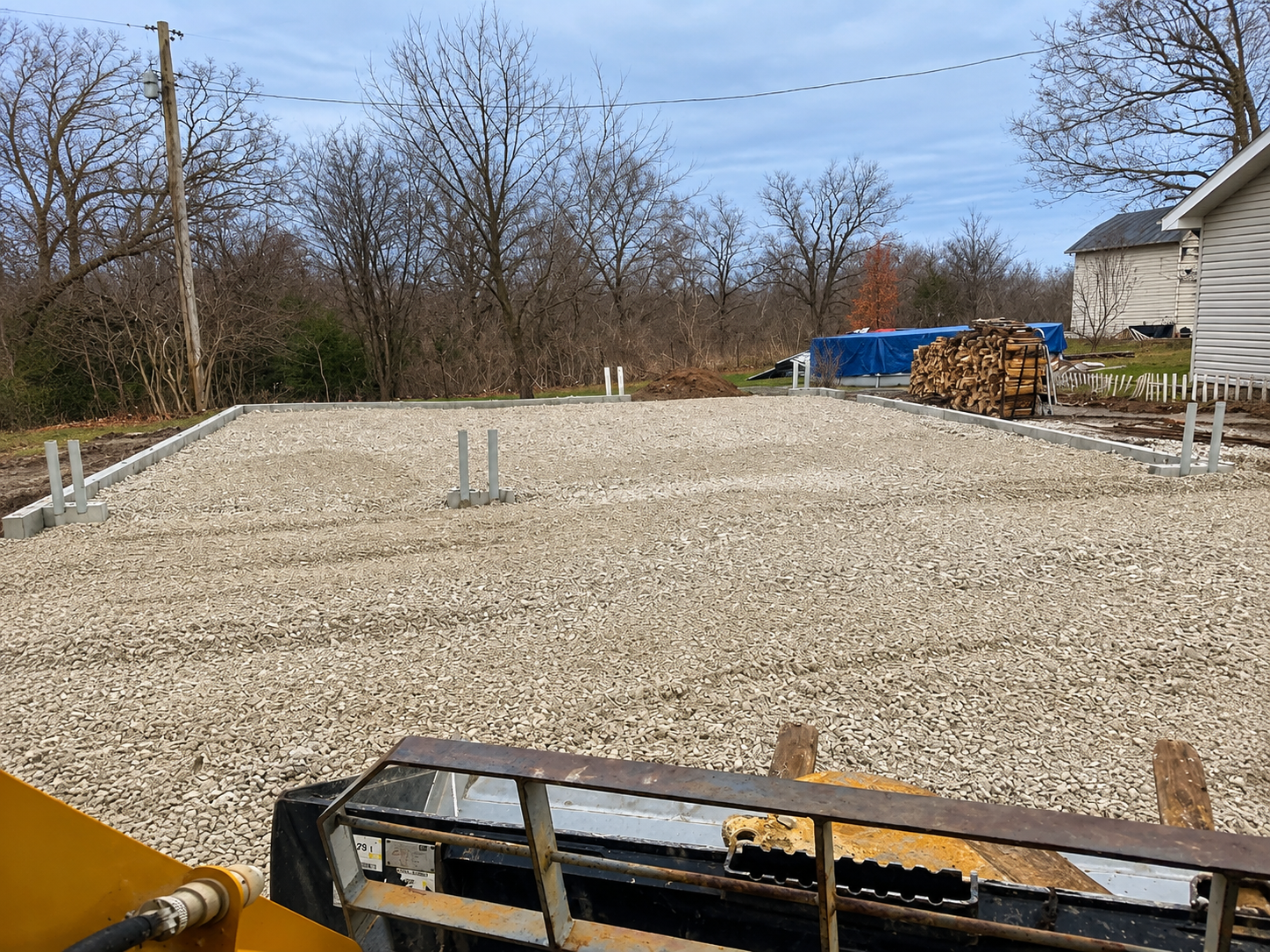 Gravel foundation with exposed pipes and a house under construction beside trees and stacked lumber