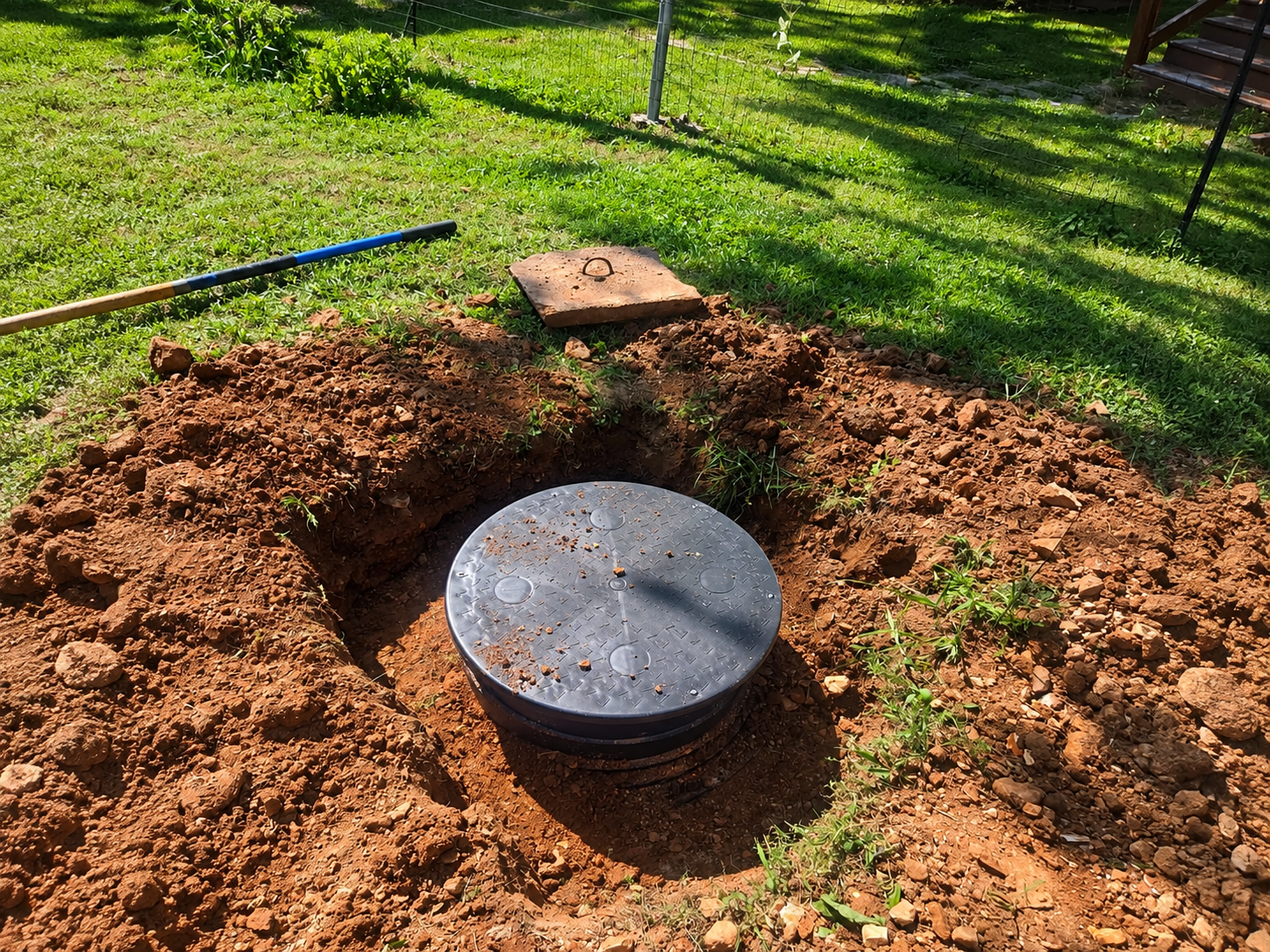 Circular concrete manhole cover in a dirt pit on a grassy yard, with a shovel nearby.