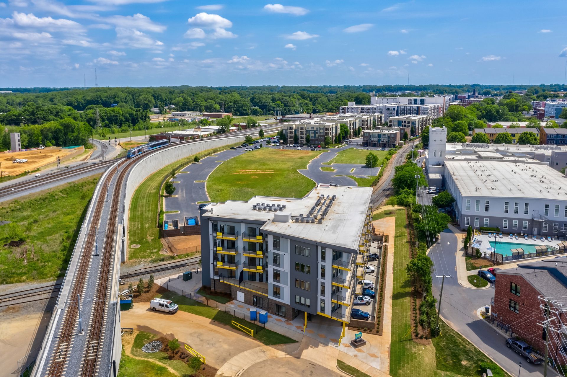 Aerial view of a modern apartment building next to train tracks and a green field on a sunny day.