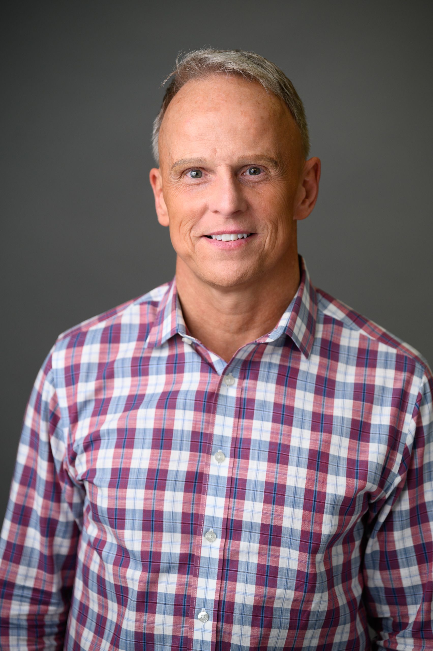 Man with short gray hair smiles, wearing a plaid shirt against a gray background.