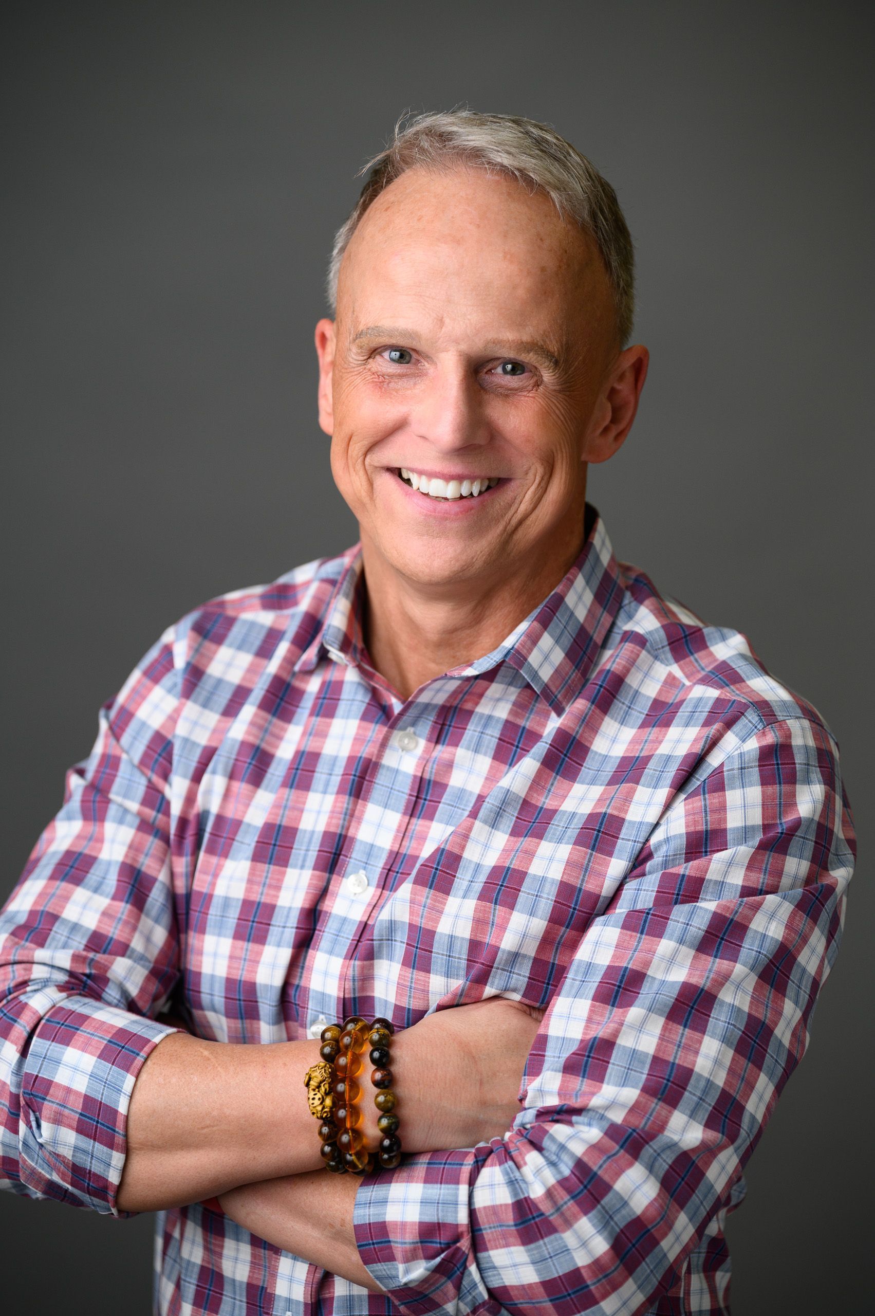 Man with arms crossed, wearing a plaid shirt, smiling, and wearing a beaded bracelet against a gray backdrop.