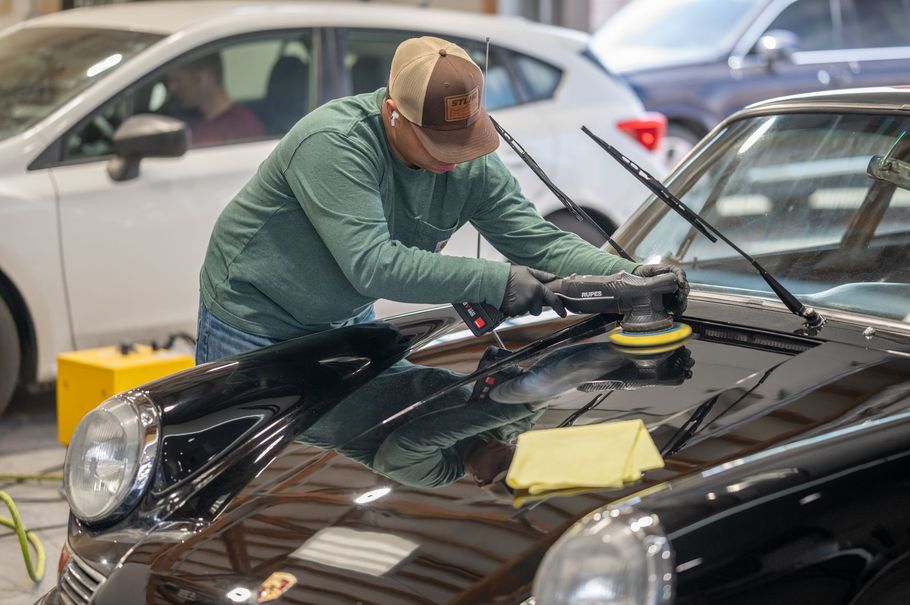 Person polishing a black car in a garage, wearing a green shirt and cap