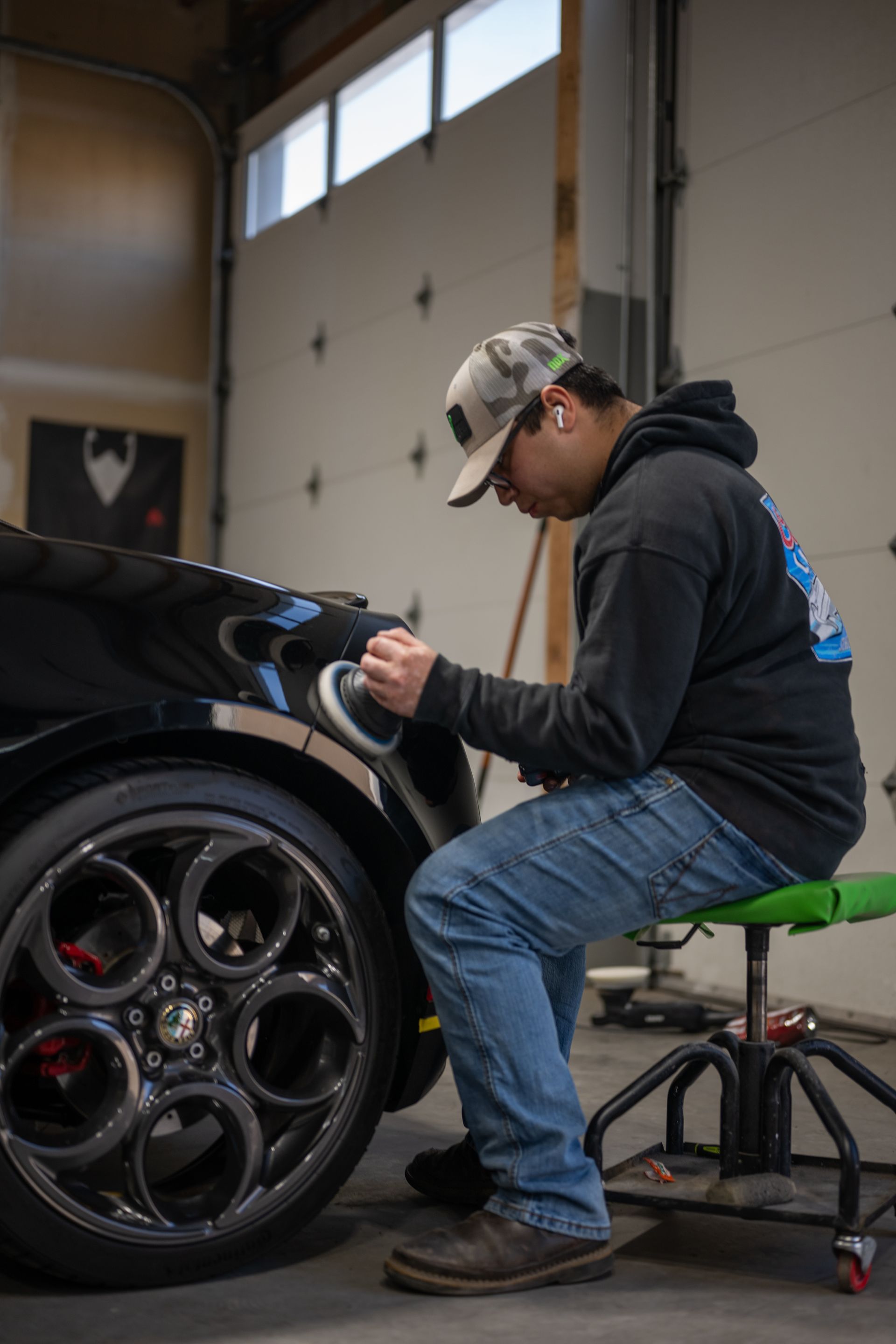Person polishing a black sports car wheel in a garage, sitting on a stool.