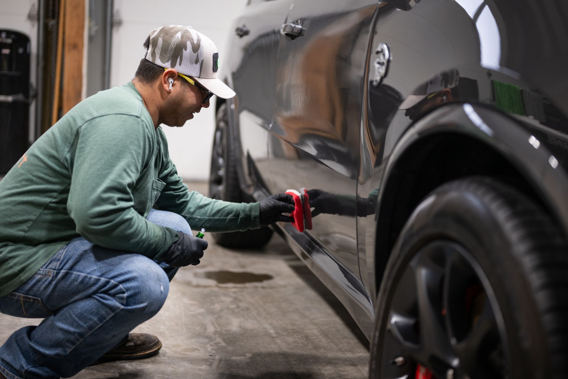 Mechanic kneeling beside a black car, polishing the side panel in a garage.