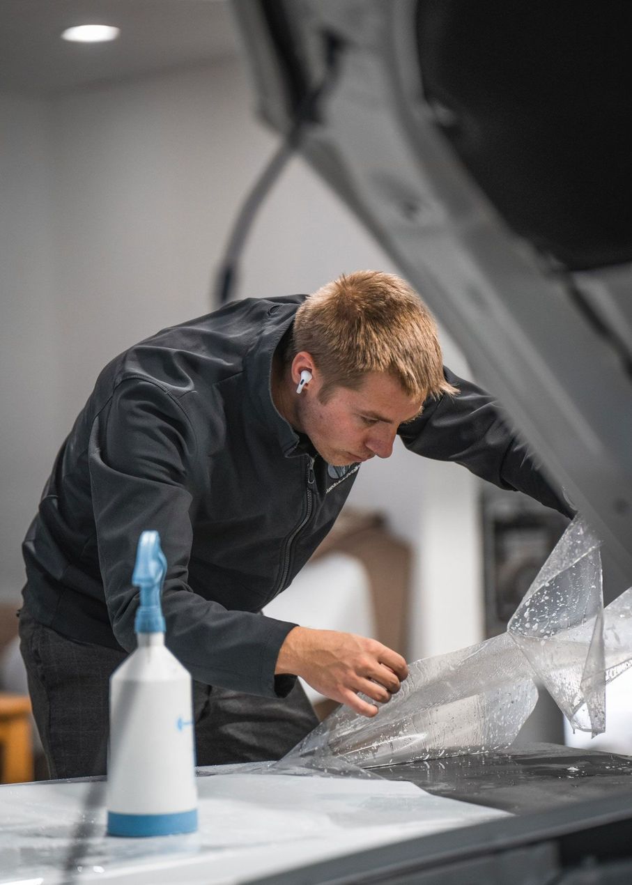 Mechanic in dark clothing inspecting the underside of a car in a garage, with a spray bottle in the foreground