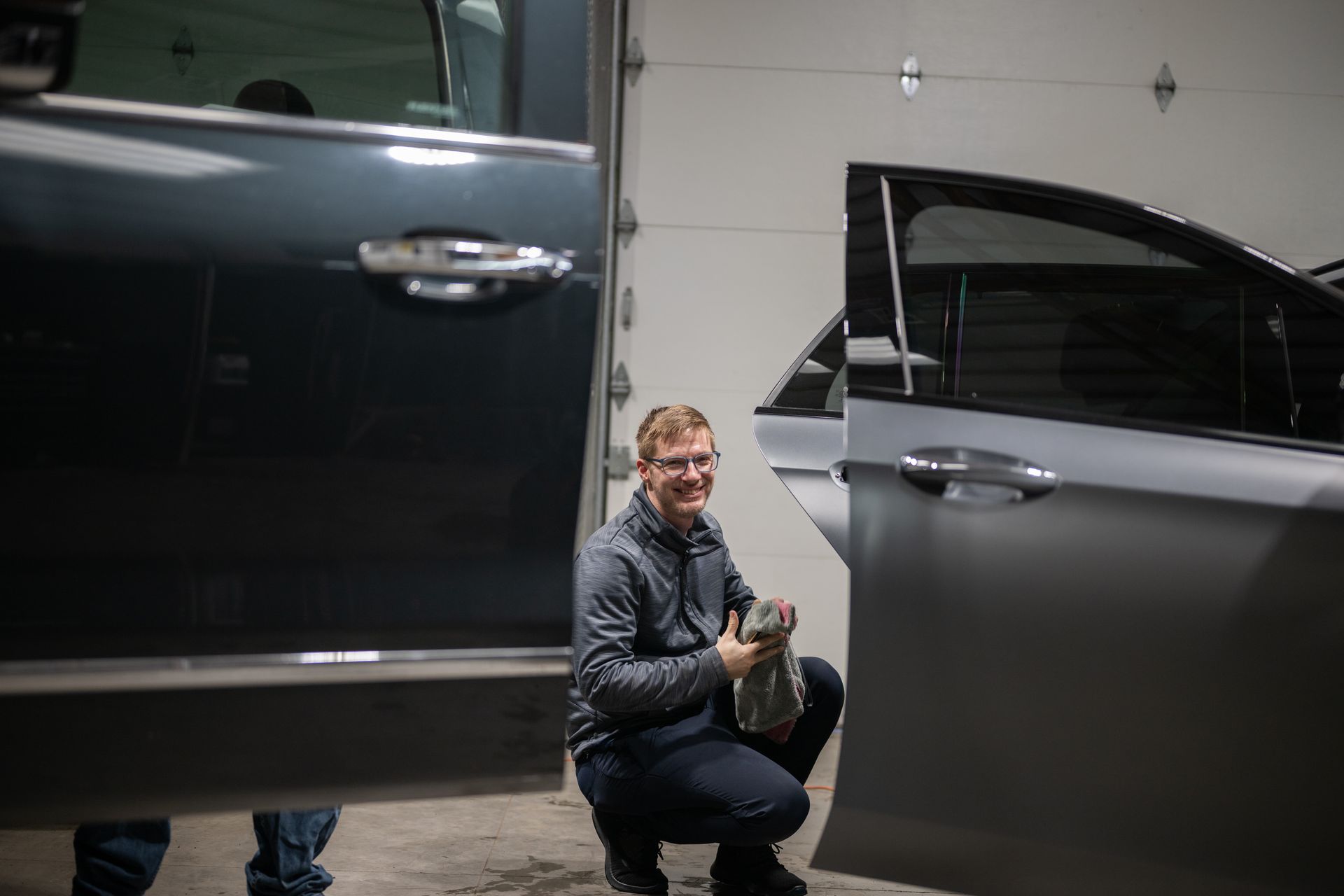 Person crouches between two dark vehicles in a garage, holding a small object.