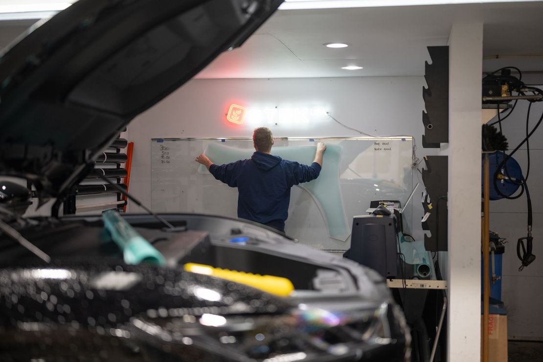 Person working on a car in a garage, arms raised toward a wall-mounted map or panel.