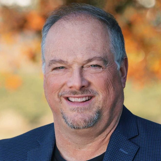Smiling man in a navy blazer against a blurred autumn background