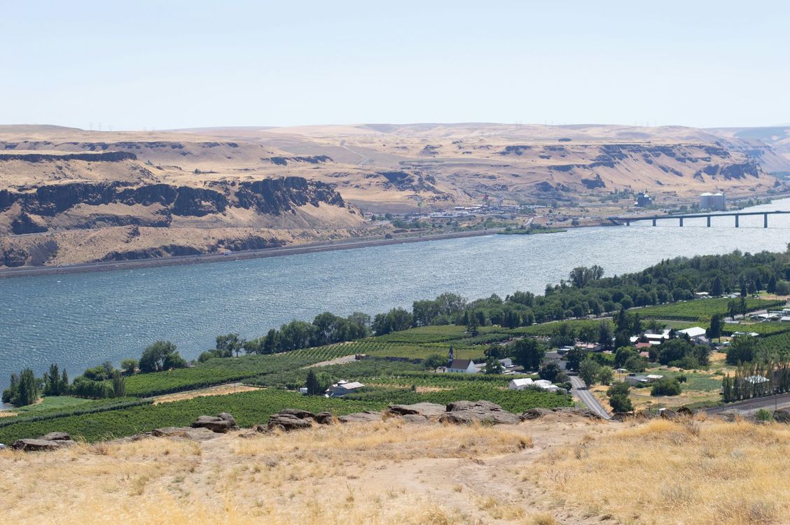 Wide river with a bridge, green fields, and dry hills under a hazy sky.