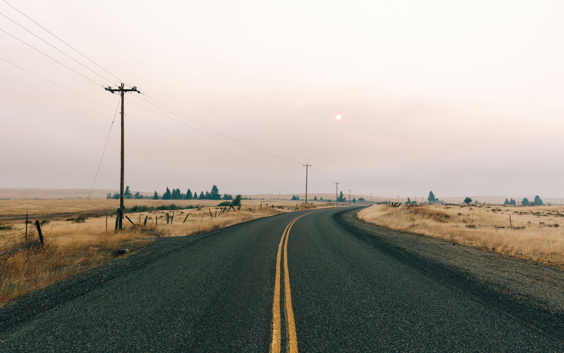 Winding rural road through dry fields at sunset, with utility poles and a hazy sky.