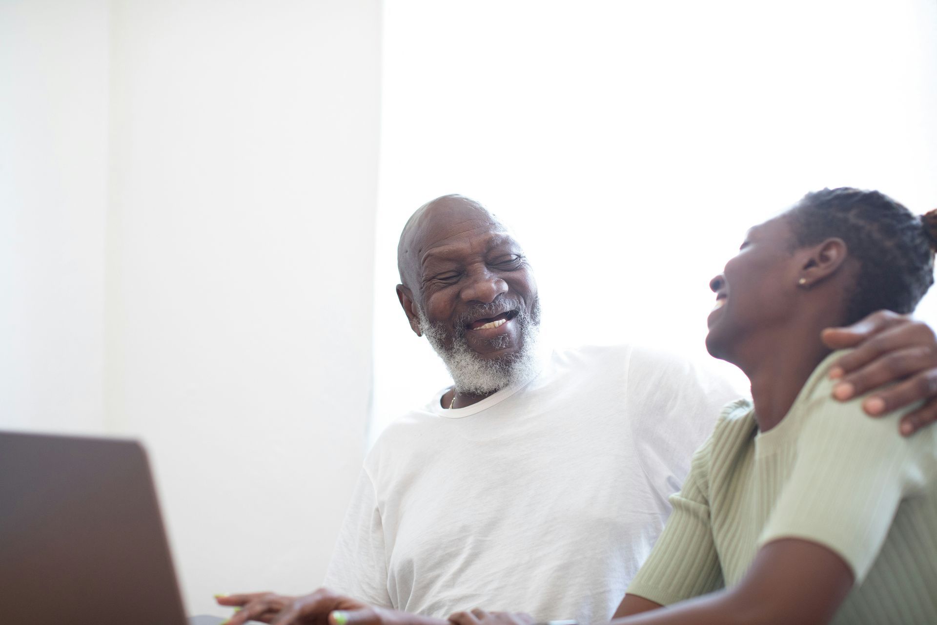 Two people smiling and talking on a couch in a bright room, with a laptop in the foreground