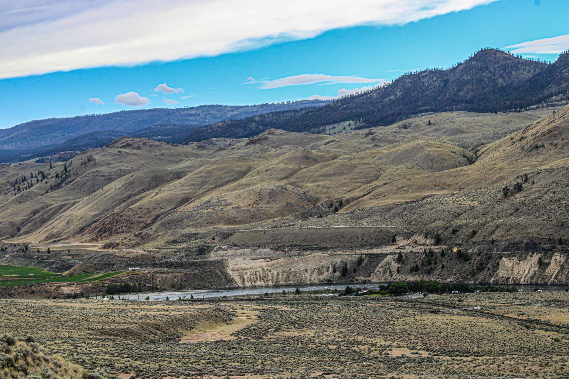 Wide arid mountain valley with rolling hills, rocky foreground, and a blue sky with clouds.