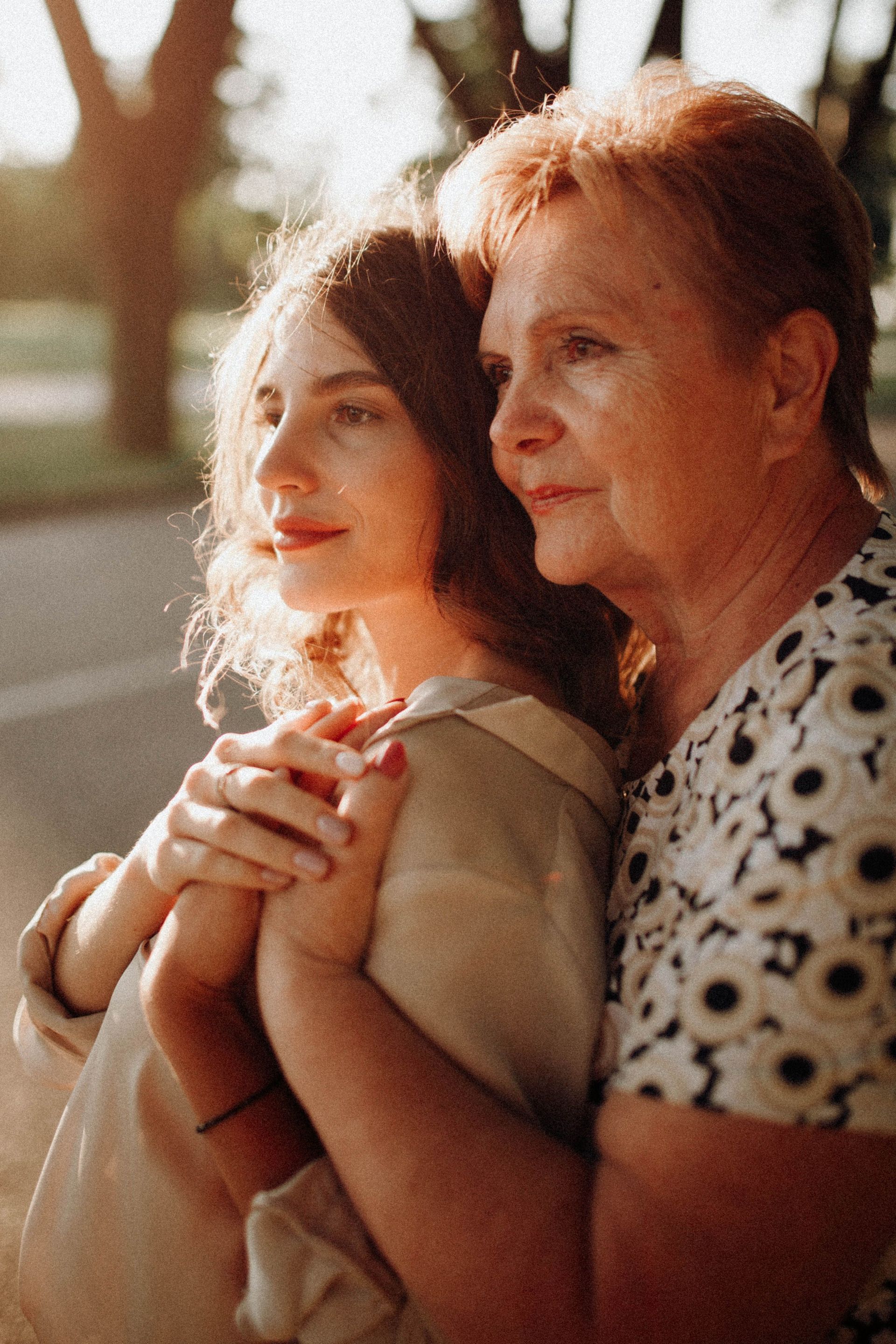 Two women standing close together outdoors in warm sunlight, holding hands and looking ahead.