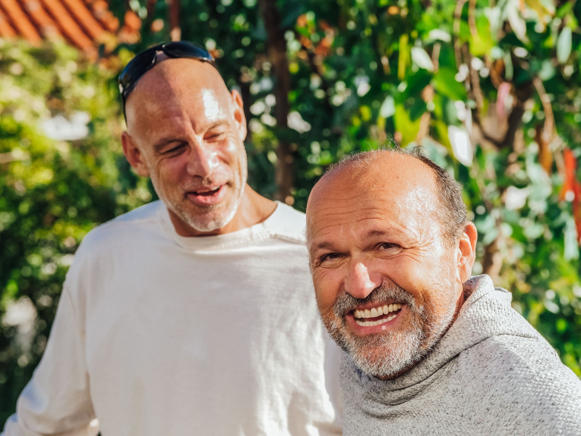 Two smiling men outdoors, one in a white shirt and one in a gray sweater, with greenery behind them.