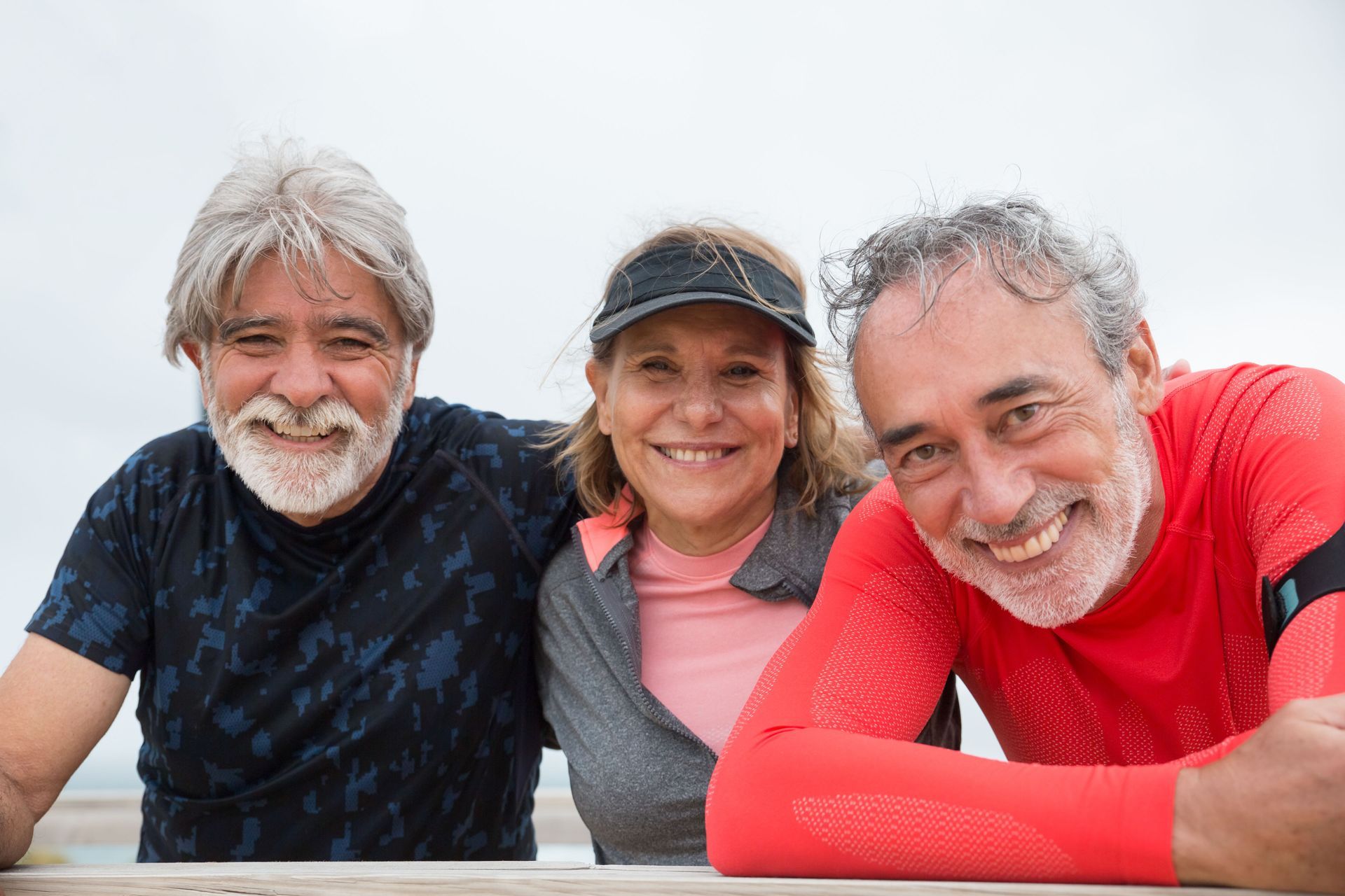 Three smiling friends leaning together at an outdoor table, wearing a black shirt, cap, and red top.