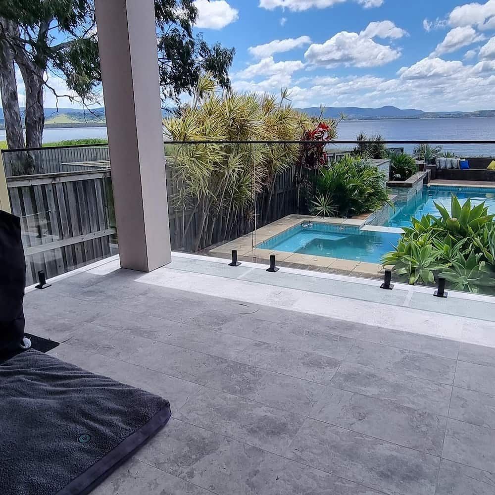 Patio overlooking pool and water. Glass railing, gray tiles, and greenery. Bright, sunny day.