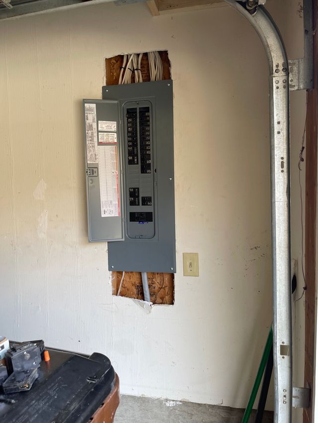 Open electrical breaker panel mounted on a garage wall with exposed wiring and unfinished drywall cutout