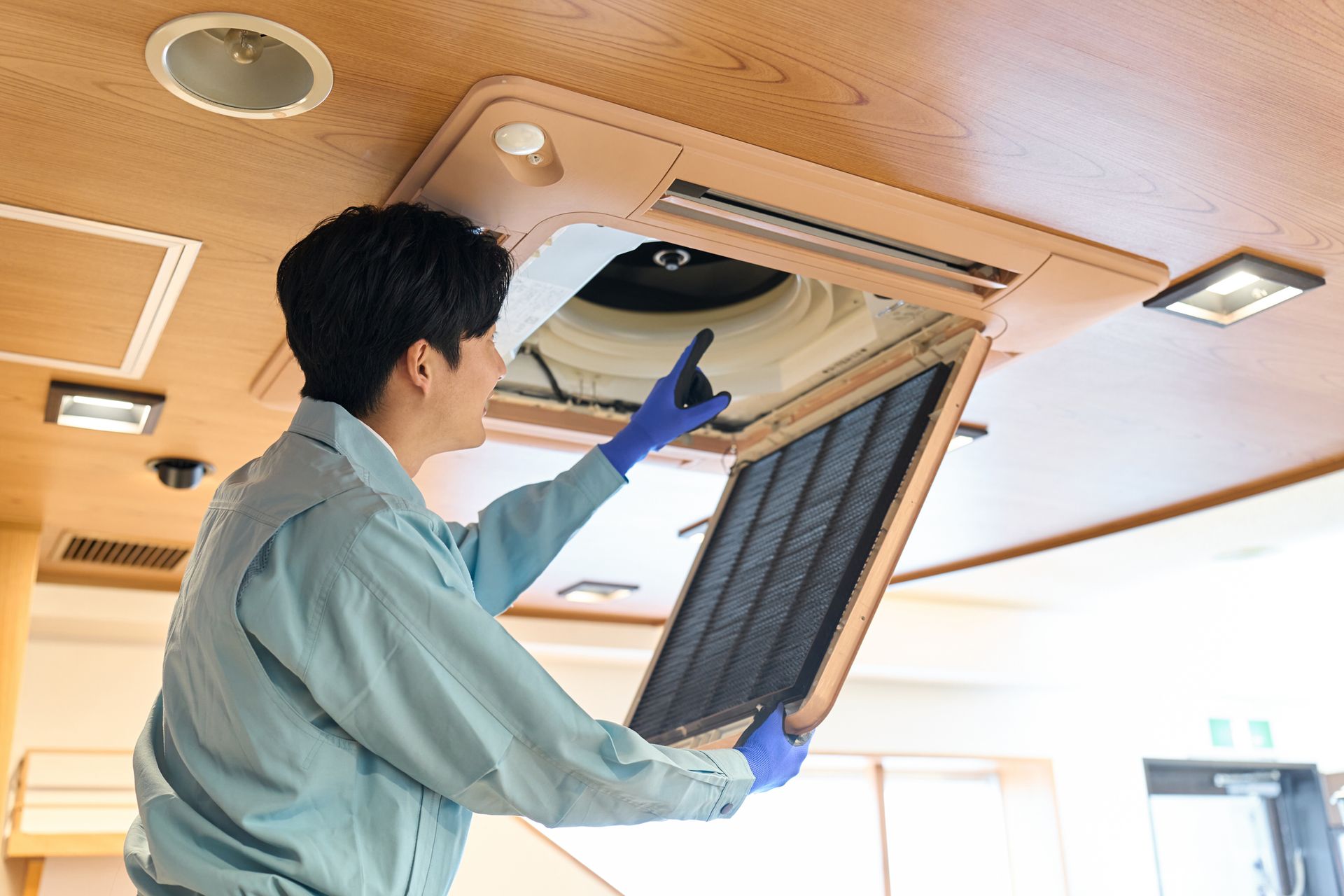 Person in blue gloves and uniform removing an air filter from a ceiling vent.