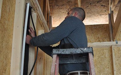 Man on a ladder installing a black cable and white pipe into a wooden wall.