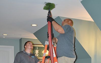 Two people cleaning a ceiling vent with a long duster on a ladder inside a room with light blue walls.