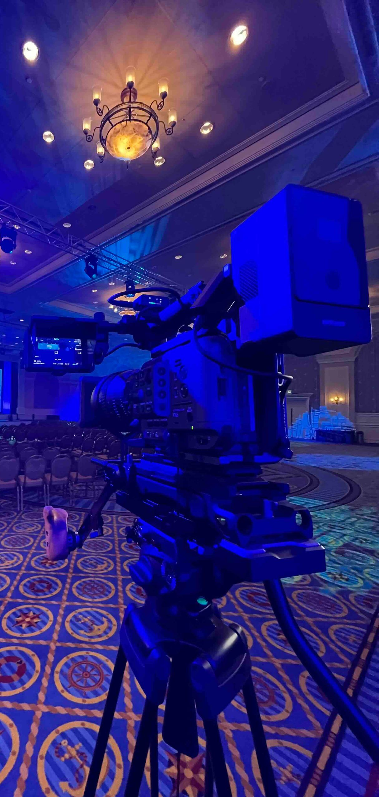 A video camera on a tripod in a dimly lit event space with a chandelier.